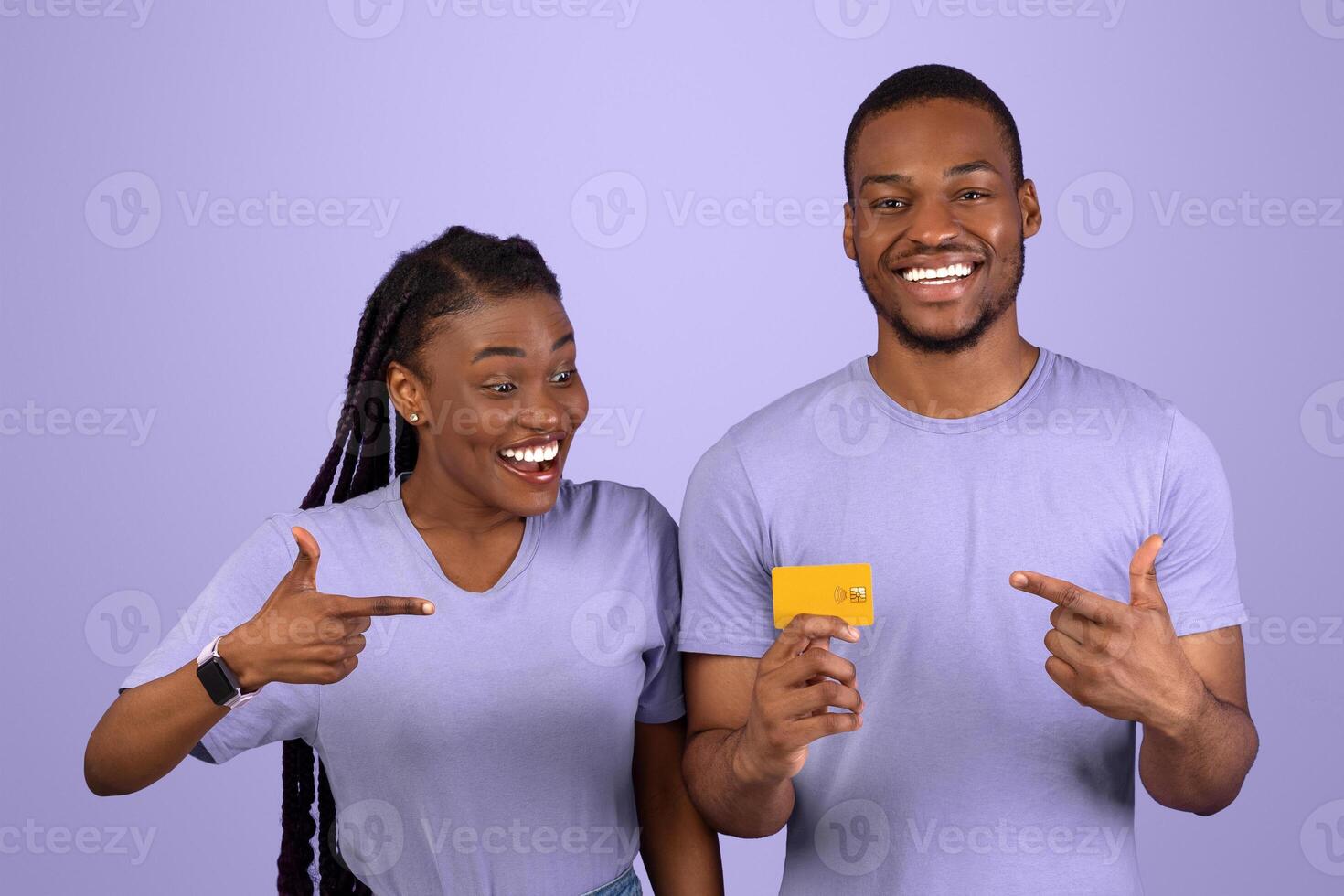Happy Black Couple Holding And Pointing Fingers At Debit Credit Card Recommending Banking Service Over Purple Violet Background. Excited Bank Customers Showing Card For Payments Posing In Studio photo