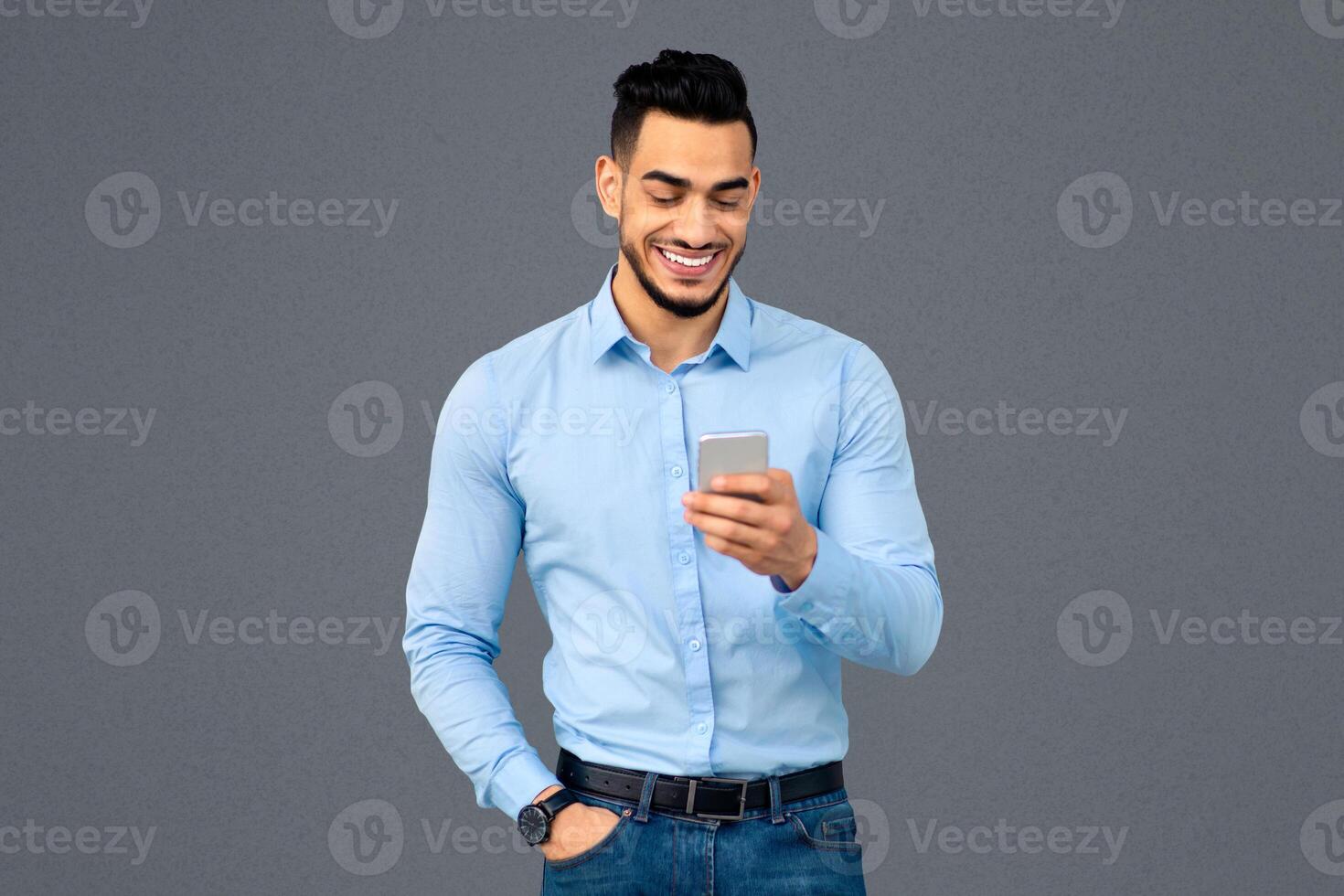 Cheerful arabic guy employee using cellphone, chatting with friends or looking for new business opportunities on Internet while having break, grey wall background, panorama with copy space photo