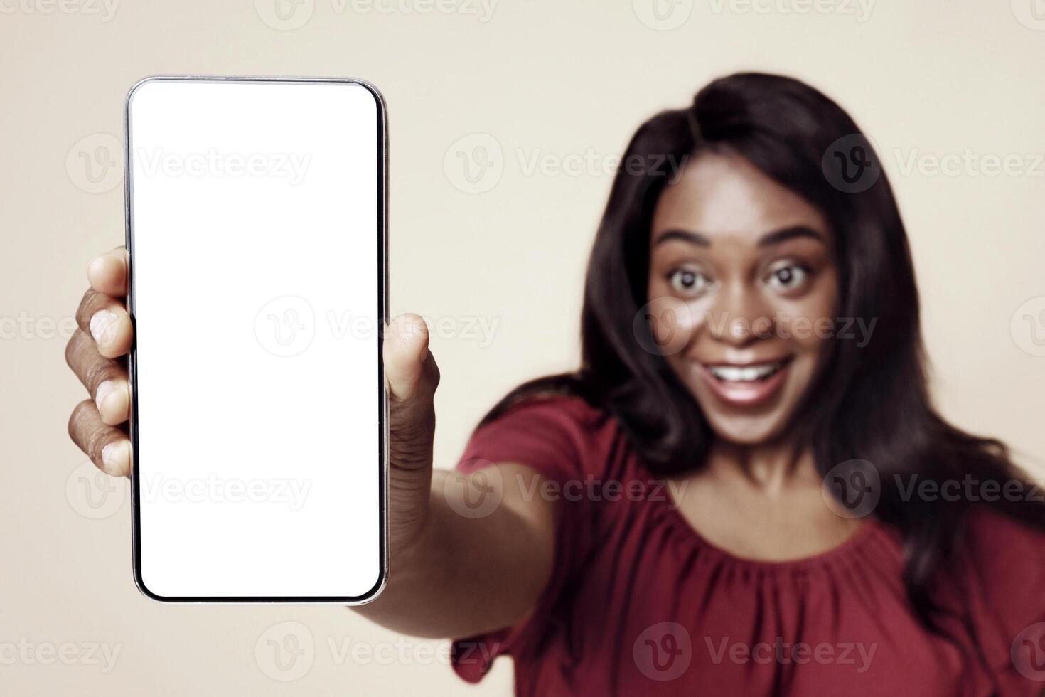 Excited african american young woman in casual outfit showing cellphone with white empty screen, recommending newest mobile application, mockup, beige studio background, copy space photo
