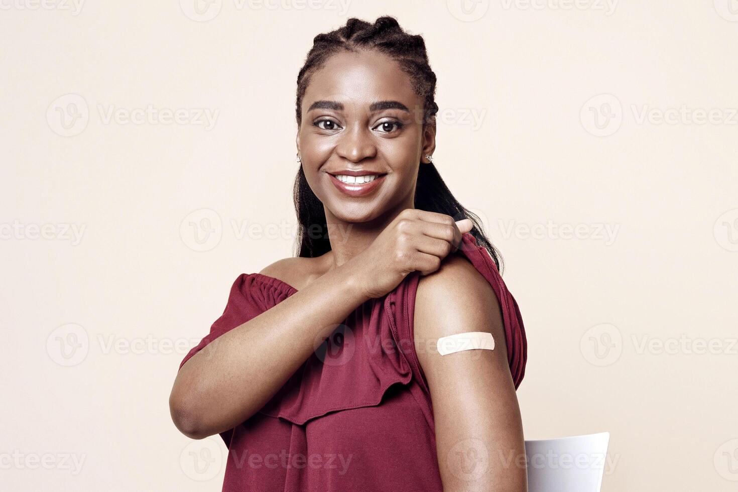Happy black lady sitting on chair, showing adhesive medical band on her shoulder after vaccination and smiling at camera, beige studio background, copy space. Immunization during COVID-19 pandemic photo