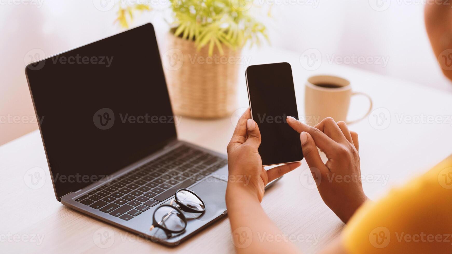 Workplace And Workspace. Over The Shoulder View Of Unrecognizable Black Lady Holding Mobile Phone With Blank Empty Screen For Mock Up Template, Sitting At Table With Computer, Plant And Coffee photo