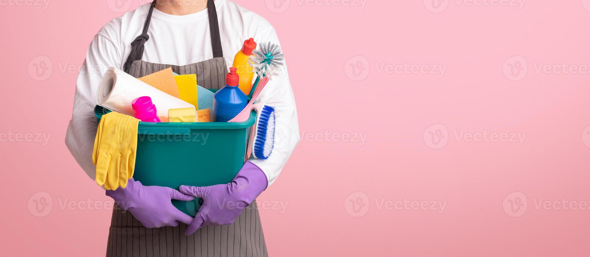 Bucket with cleaning supplies and tools in hands of unrecognizable man in apron standing on pink background, cropped image, panorana with free space photo