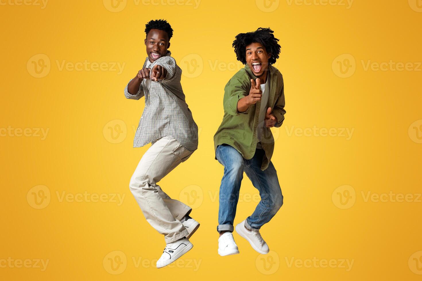 Two Cheerful Black Guys Pointing At Camera While Jumping Over Beige Studio Background, Emotional Young African American Males Saying Gotcha, Having Fun Together, Full Length Shot, Copy Space photo