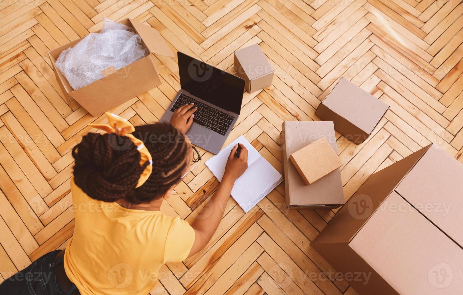 Above top high angle view of young african american woman typing on laptop keyboard, writing in notebook, lying on the floor near cardboard boxes. Computer with blank empty screen for mockup photo