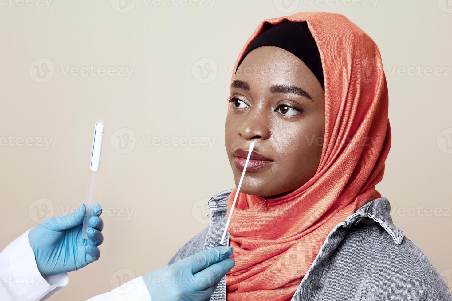 Coronavirus, COVID-19 pcr test concept. Medical worker taking nasal swab for coronavirus sample test from potentially infected muslim african american woman, beige studio background, closeup photo