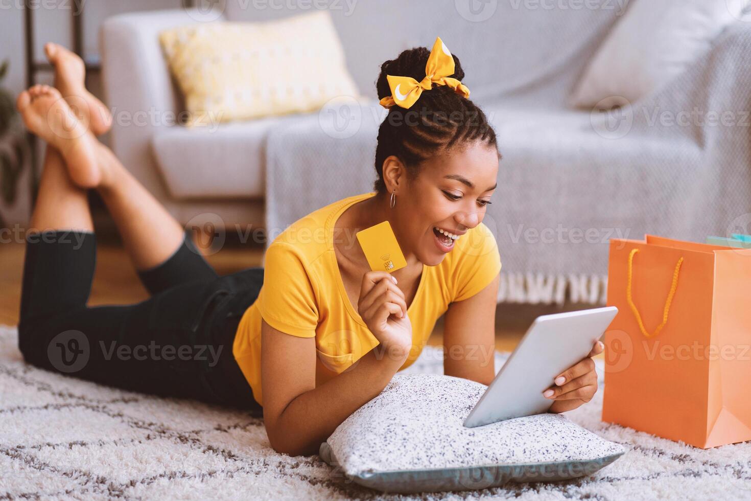 Excited african girl showing credit card, using tablet, lying on the floor photo