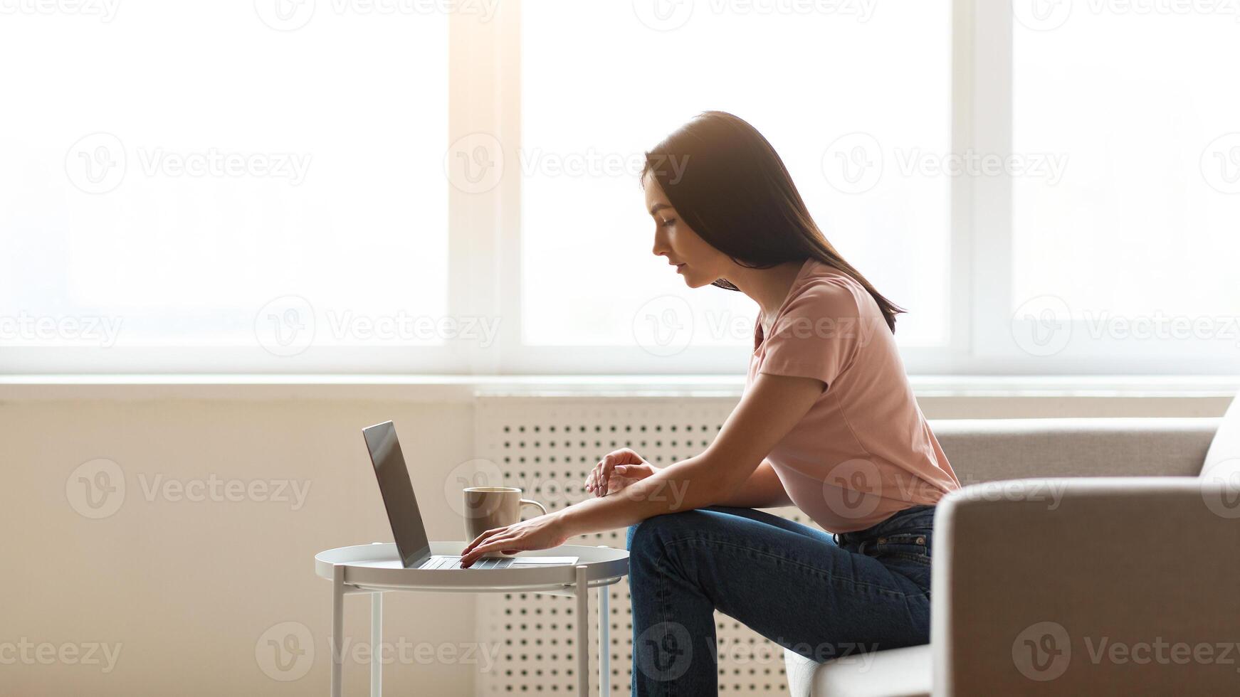 Female Freelancer Working On Laptop Computer Sitting On Sofa Near Window At Home. Modern Lifestyle And Distant Career. Side-View photo