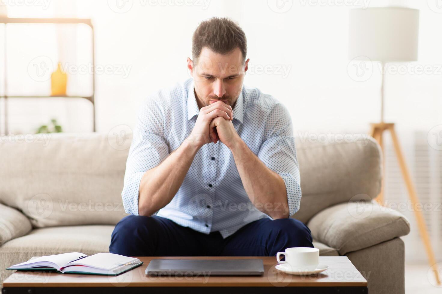 What To Do. Contemplating serious adult man sitting on couch alone home, looking at desk and thinking, copy space photo