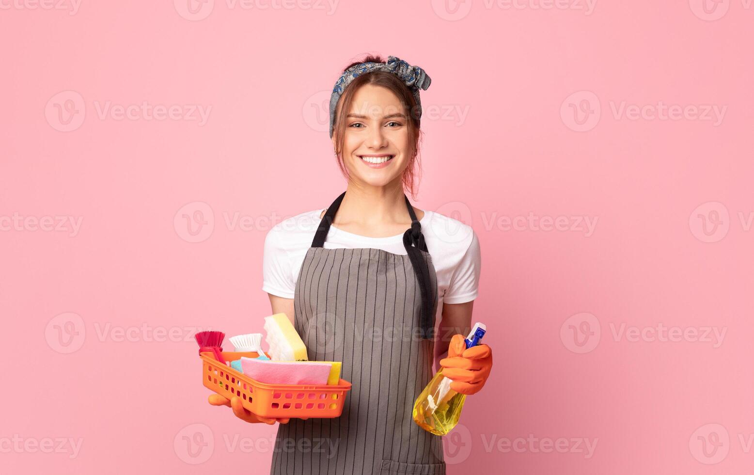 Happy Housewife In Apron Holding Detergent Spray And Cleaning Tools, Looking At Camera While Posing Over Pink Background With Empty Space photo