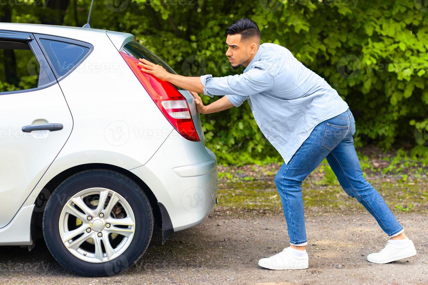 Arab guy in stylish casual outfit pushing broken car, side view. Middle-eastern man travelling with friends or lover, got stuck on the road to countryside, pushing automobile hatchback photo