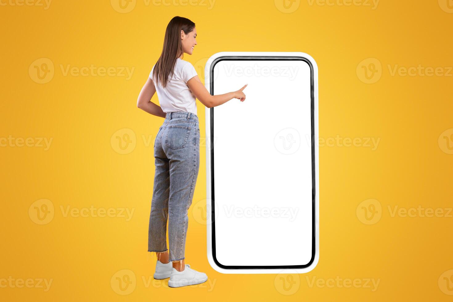 Full Body Length Back View Of Woman Using Big Smartphone With Blank White Screen And Touching Display Panel With Finger, Cheerful Lady Standing On Yellow Background, Ordering Food Delivery, Mock Up photo