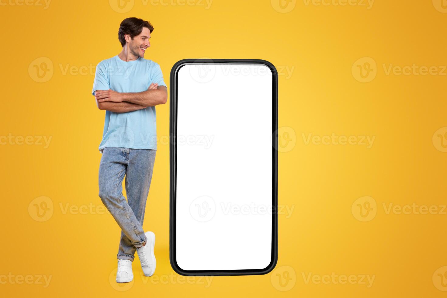 Cheerful smiling casual guy standing with crossed hands looking at giant big cell phone, empty white screen template, orange studio wall. Mock up for app or website, free copy space, full body length photo