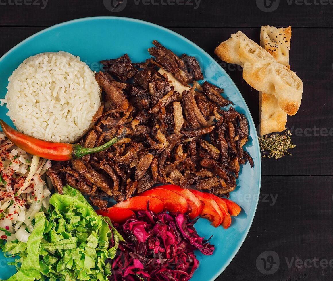 Delicious iskender kebab is served on a blue plate with rice, fresh vegetables and pita bread close up photo