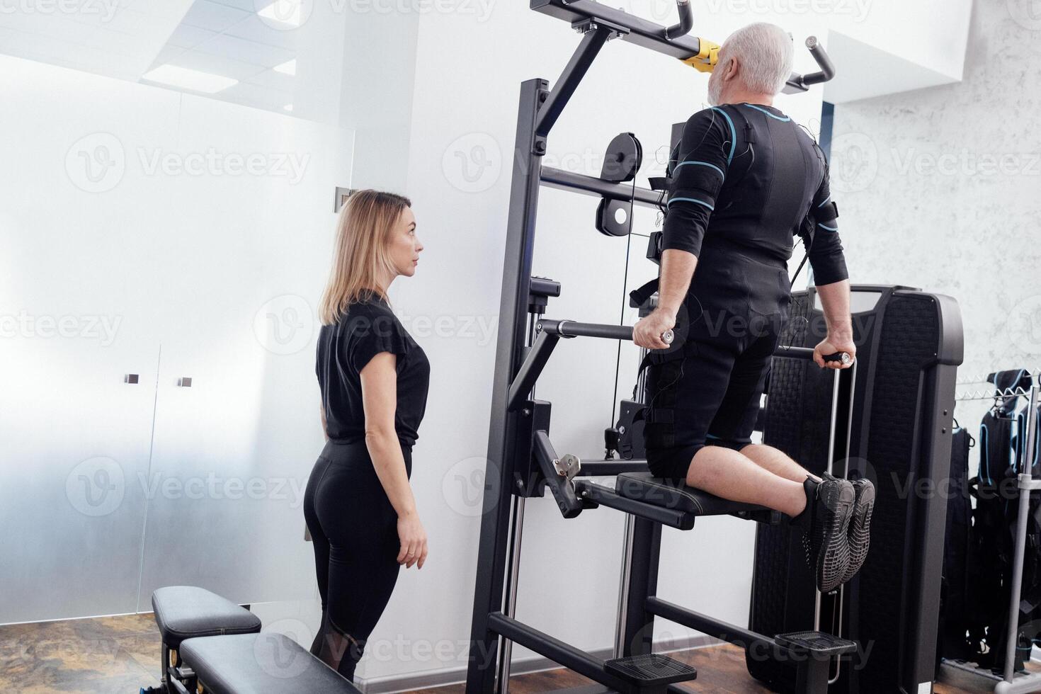 Elderly greyhaired man in ems suit is engaged on the uneven bars on the gravitron simulator with personal trainers in gym. photo