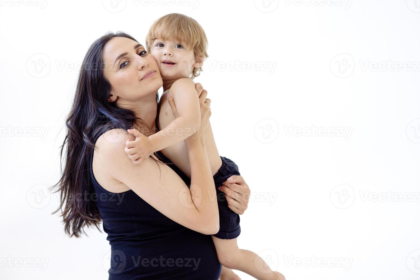 A smiling pregnant woman in a black dress hugs her baby. A charming young mom gently touches her son. Isolated white background. The embrace of a mother and her little boy. photo