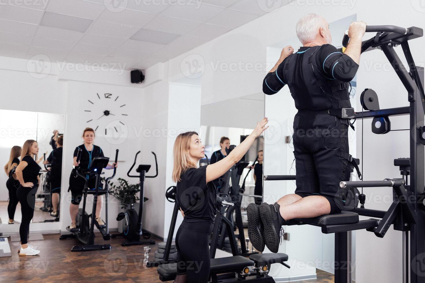 Elderly couple of people in ems suits are engaged in sports equipment with personal trainers in gym. photo