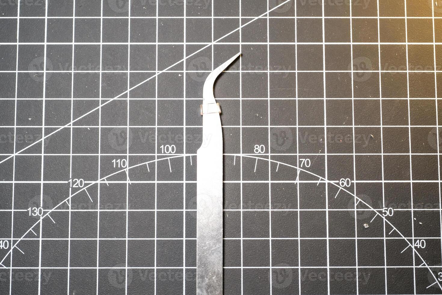 Metal precision tweezer with pointed tip. Tweezer is on a black cutting mat with lines. A tool that functions to clamp and pull out small objects. Negative Space. Beauty and grooming tools photo