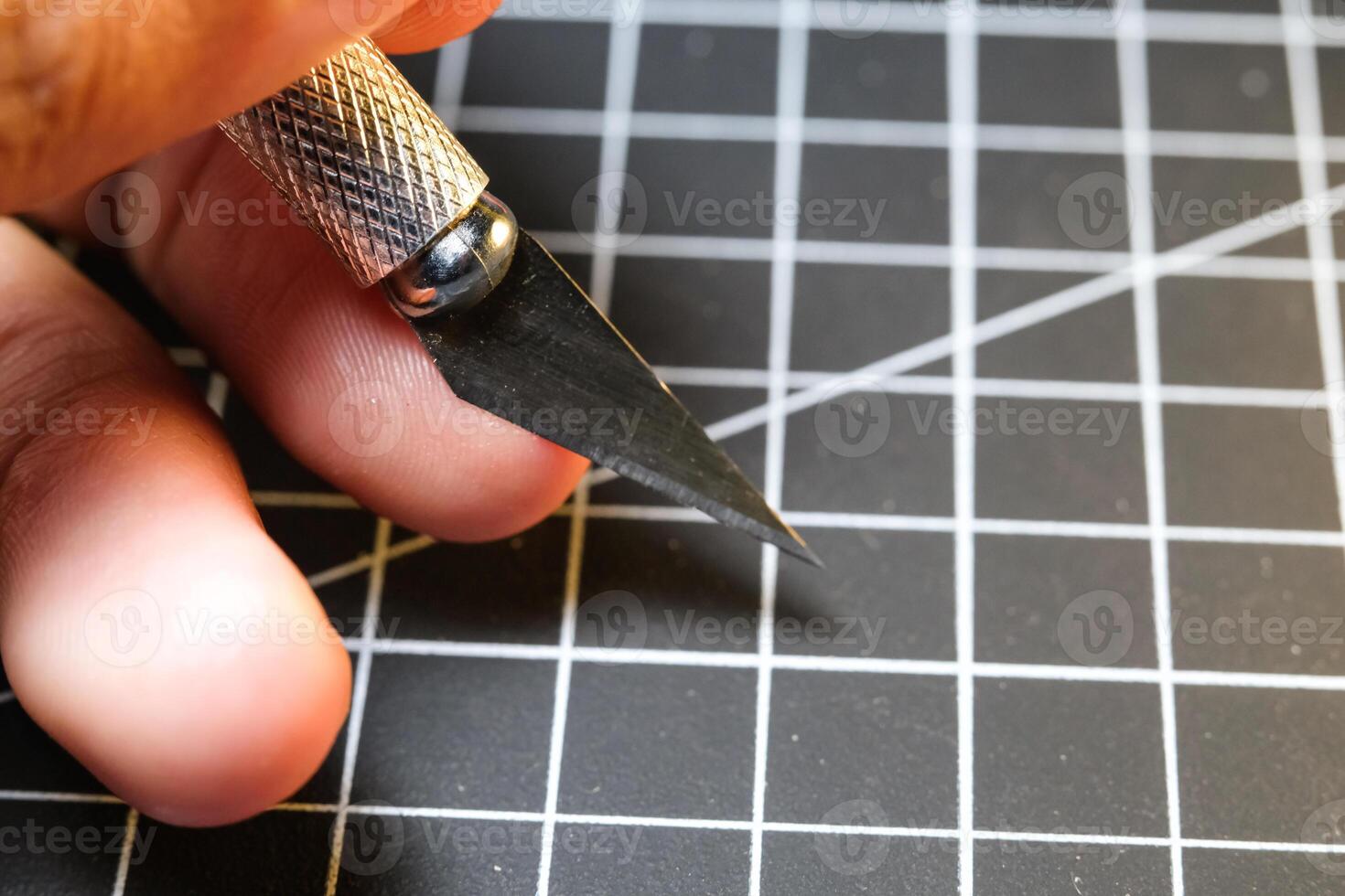 Closeup of a hand holding a cutting pen on black striped rubber mat. Sharp cutting blade used to cut plastic parts. Metal tools and equipment. Tools for hobbyist robot model kit assembly. photo