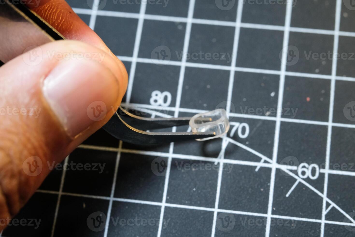 A man's hand on black cutting mat holds a Tweezer for pinching tiny parts. Black Cutting mat background with white lines. The concept of a master model kits hobby. Negative space photo