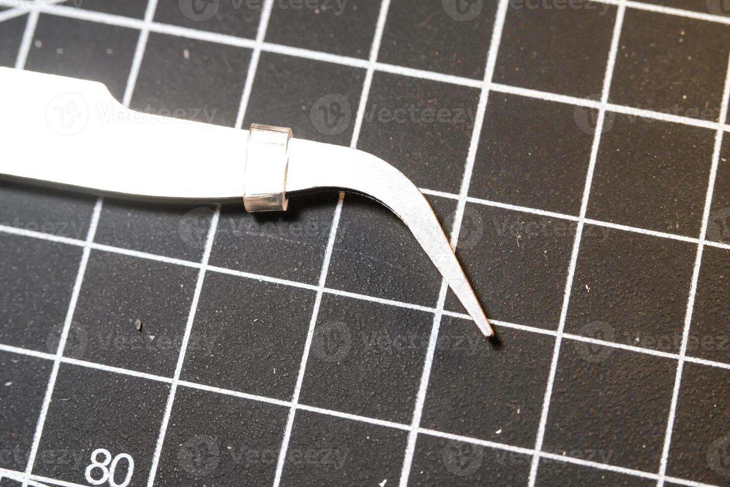 Metal precision tweezer with pointed tip. Tweezer is on a black cutting mat with lines. A tool that functions to clamp and pull out small objects. Negative Space. photo