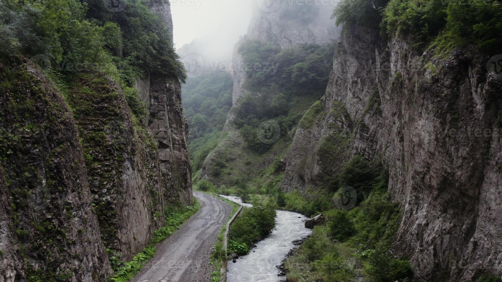 Russia, Caucasus. Vertical panning from a quadcopter. A narrow mountain road and a bubbling river in the lowlands between sheer rocky mountains covered with green vegetation and clouds photo