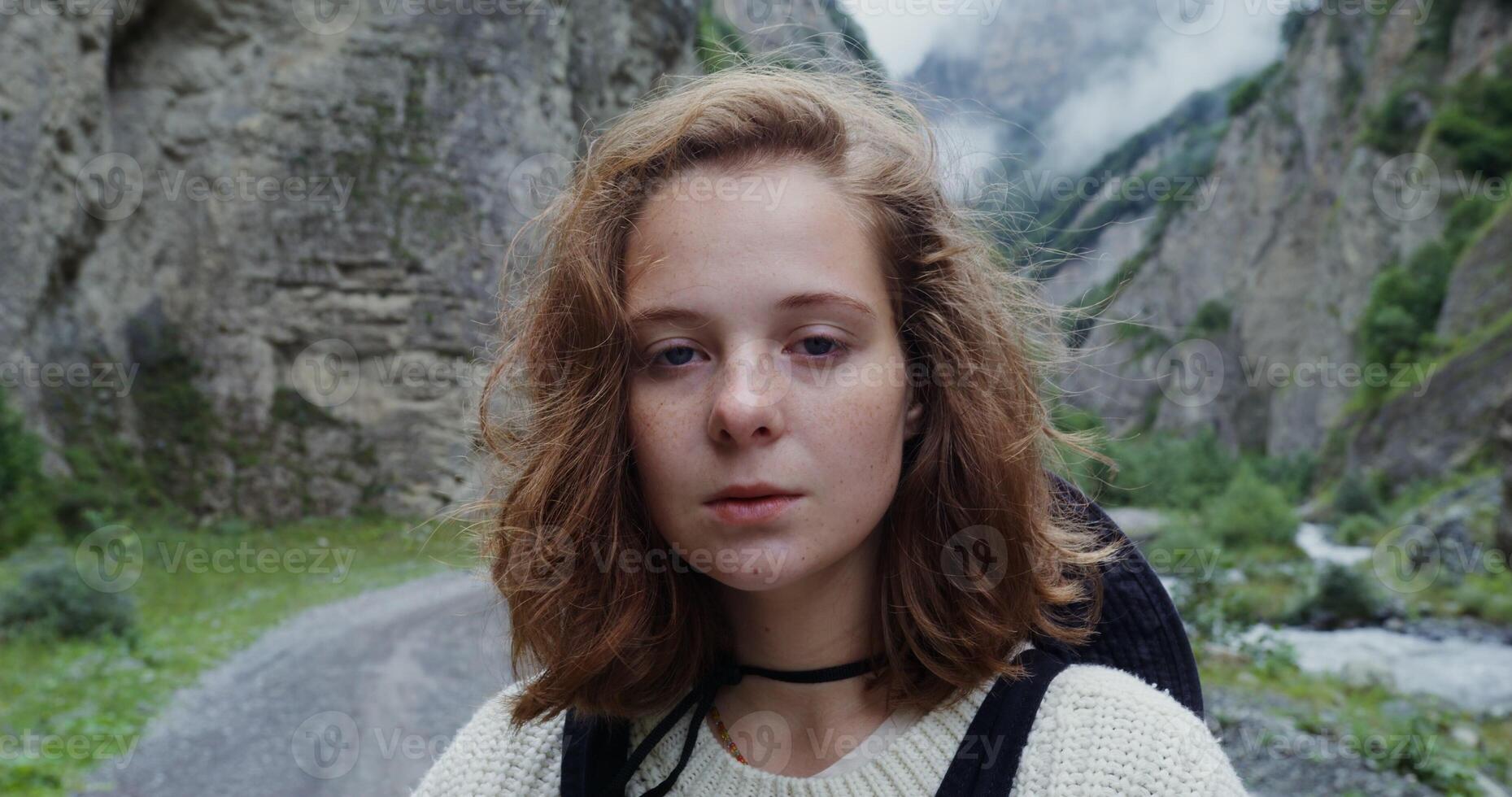 Russia, Caucasus, Elbrus. Young red-haired girl of European appearance with freckles smiling while looking at the camera, standing on a mountain road among the rocks in the daylight photo