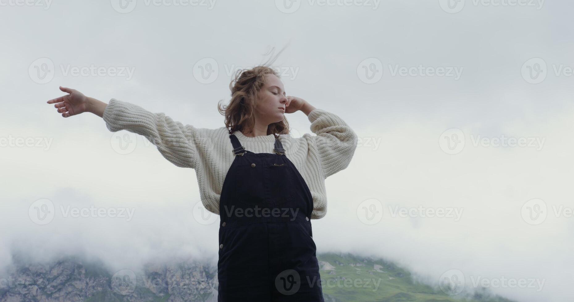 Russia, Caucasus. A beautiful young woman of European appearance, dressed in a warm sweater, stands on the parapet against the background of green hills and makes smooth movements with her hands photo