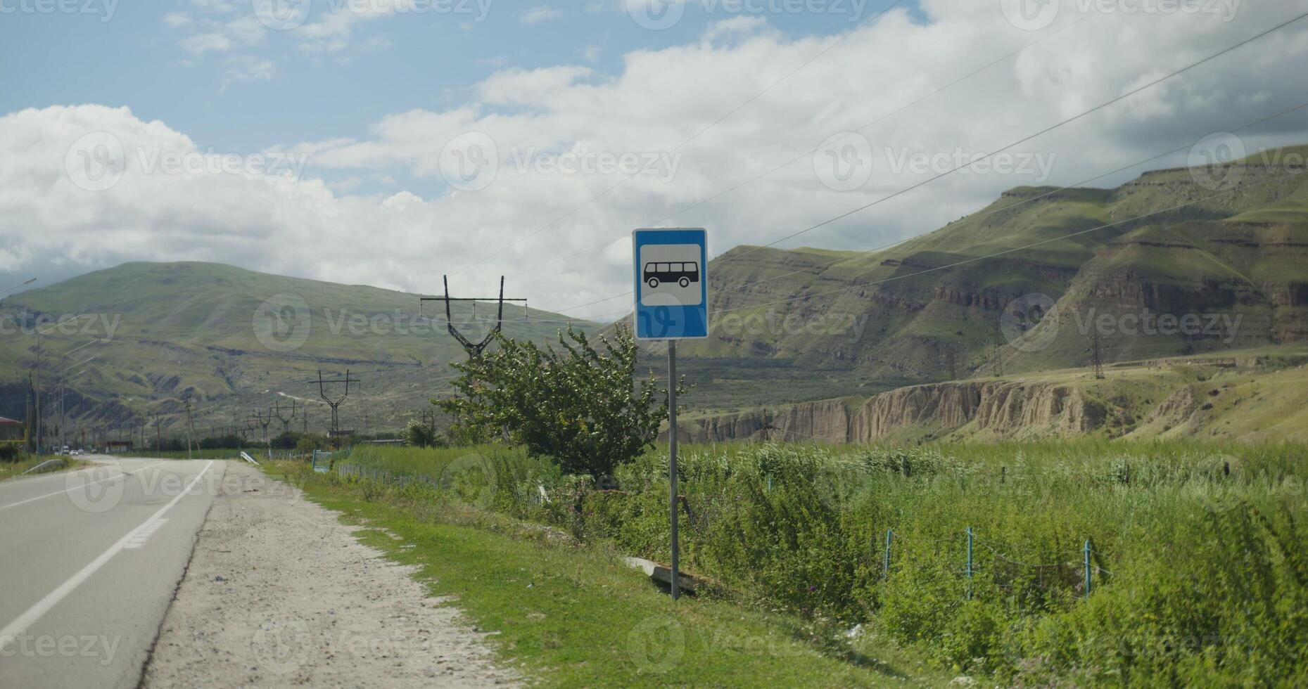 View of the bus stop road sign against the backdrop of rocky mountains, the tops of which are covered with thick white clouds. Vertical photo