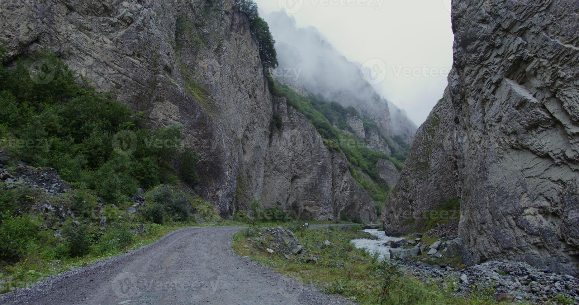 A lone car drives along a mountain road among sheer cliffs covered with sparse vegetation. The tops of the mountains are covered with clouds. Daylight photo