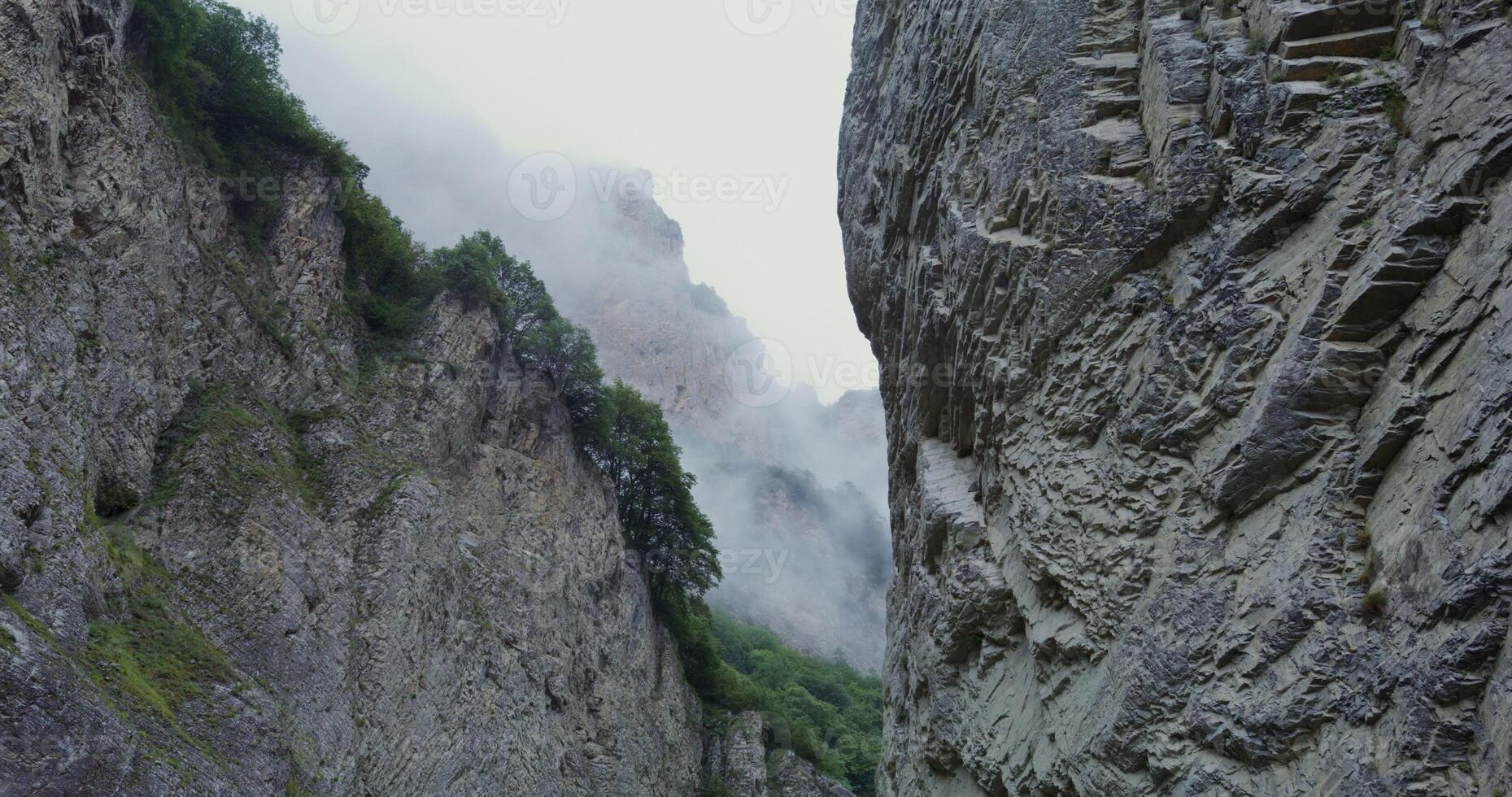 A deep crevice between sheer rocky cliffs covered with sparse vegetation. The tops of the mountains are covered with clouds photo