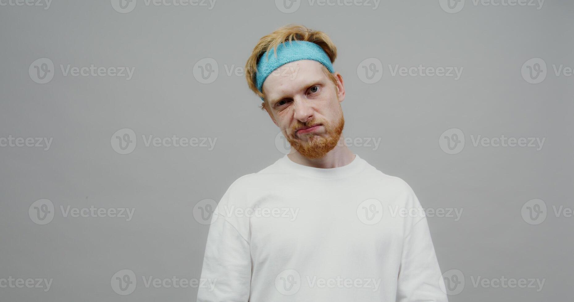 A red-haired guy with a beard and a sports band on his head, looking bored, trains his biceps with small dumbbells. Shooting on a light solid background. photo