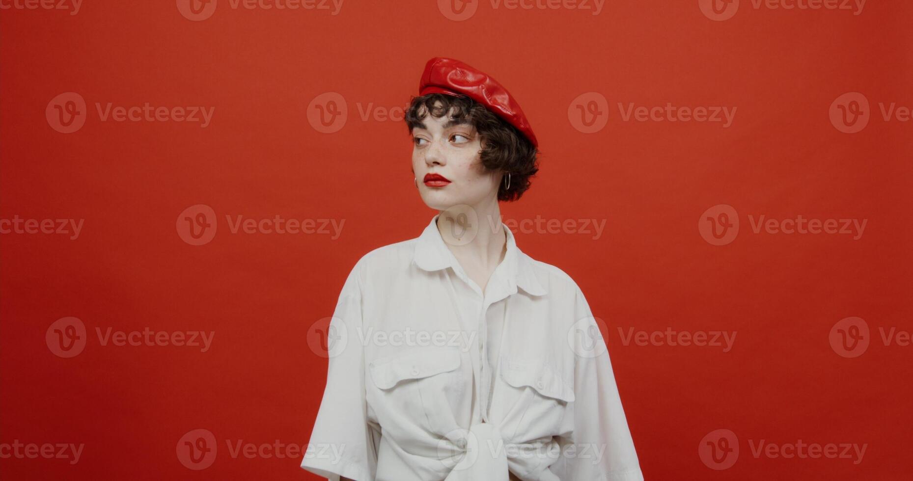 A brunette woman of European appearance with a short haircut in a red panama hat flirts, playfully straightening her hair. Shooting on a red background. photo