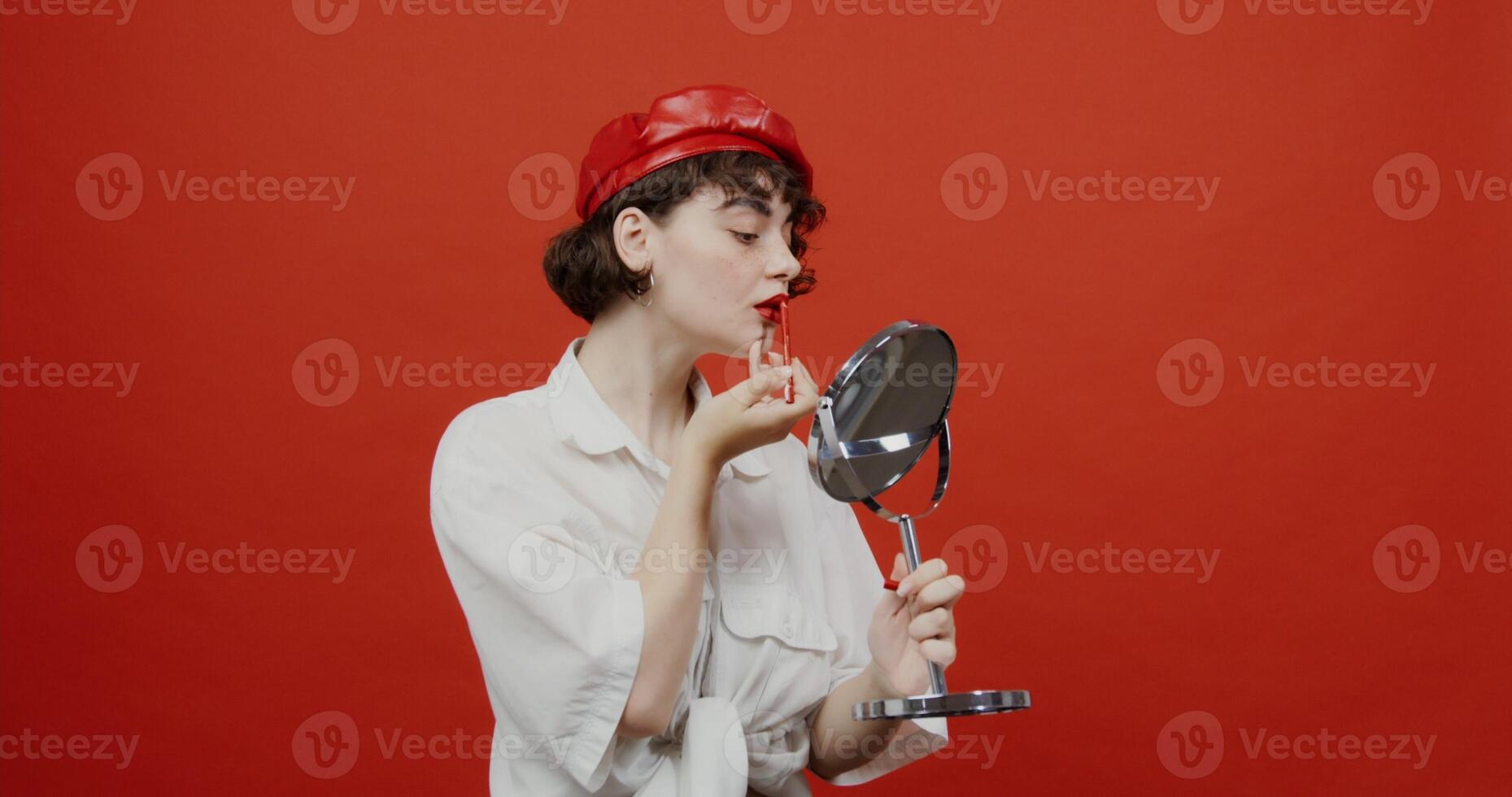 A woman of European appearance with a short haircut in a red panama hat paints her lips with a red pencil, holding a mirror in her hand. Shooting on a red background. photo