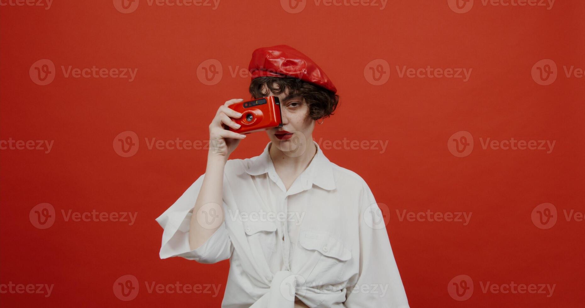 A brunette woman of European appearance with a short haircut in a red panama hat makes a photo on a red camera and laughs. Shooting on a red background.