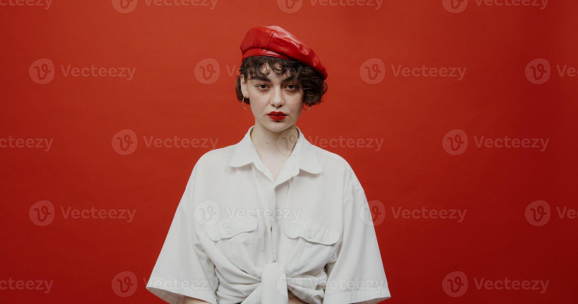 A brunette woman of European appearance with a short haircut in a red panama hat looks at the camera with a bored look. Shooting on a red background. photo