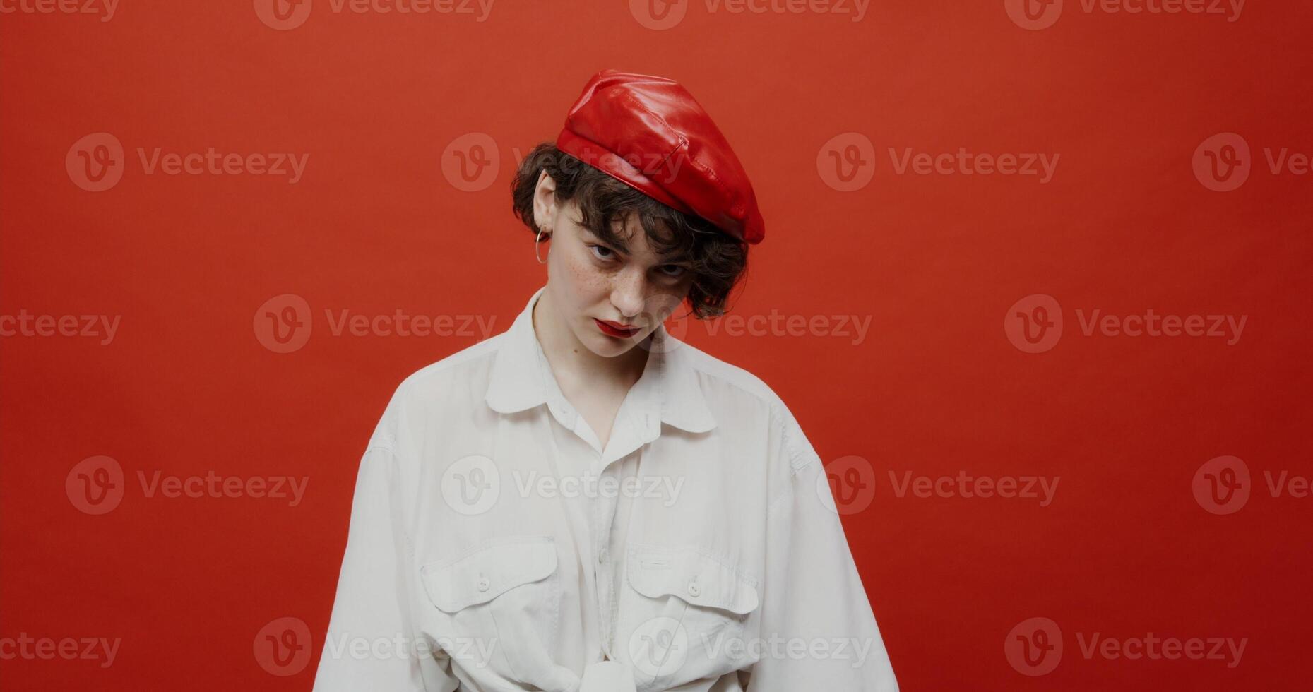 A brunette woman of European appearance with a short haircut in a red panama hat looks angrily from under her forehead with a cruel look. Shooting on a red background. photo