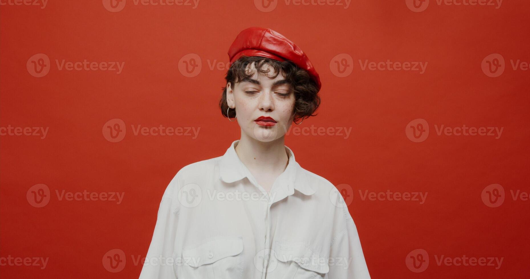 A brunette woman of European appearance with a short haircut in a red panama hat is frightened by something from the side, bends over and covers her face with her hands. Shooting on a red background. photo