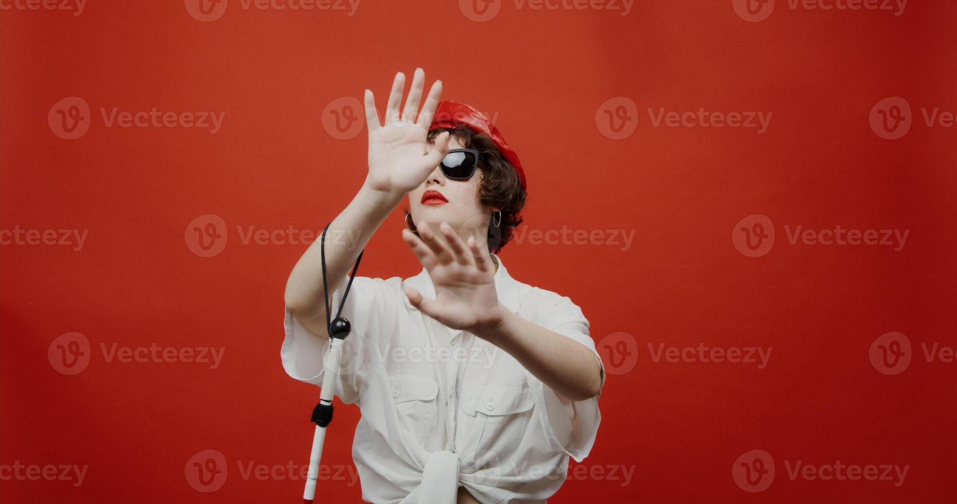 A blind woman of brunette European appearance with a short haircut in a red panama hat and sunglasses with a cane on her hand is trying to grope for something ahead. Shooting on a red background. photo