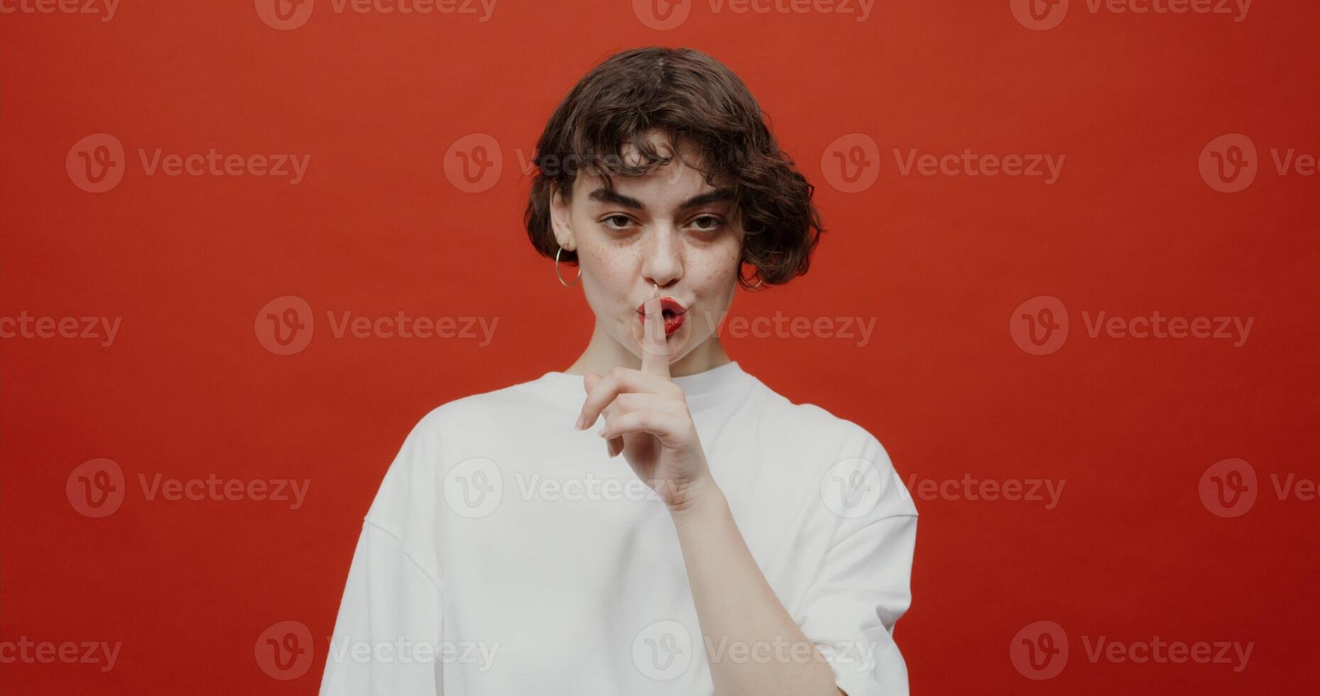 A brunette woman of European appearance with a short haircut brings her index finger to her mouth. makes a sign to be silent. Shooting on a red background. photo