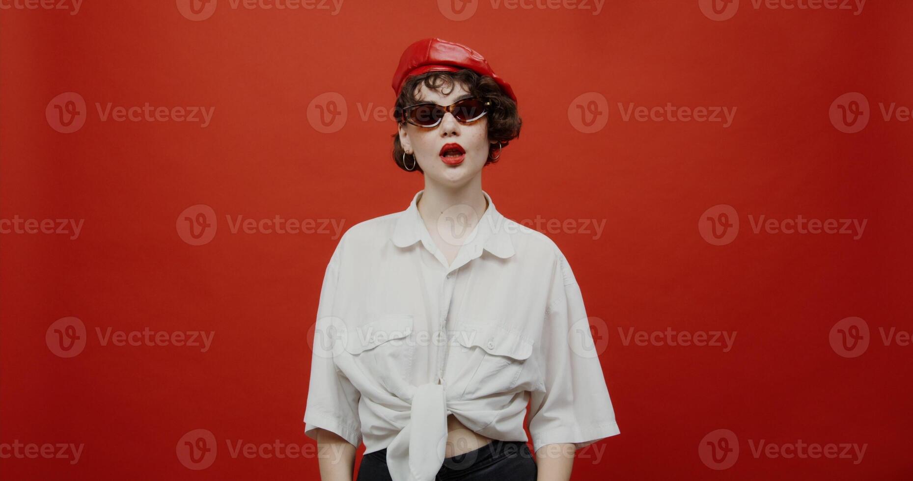 A brunette woman of European appearance with a short haircut in a red panama hat and sunglasses is flirting, looking playfully from under her glasses. Shooting on a red background. photo