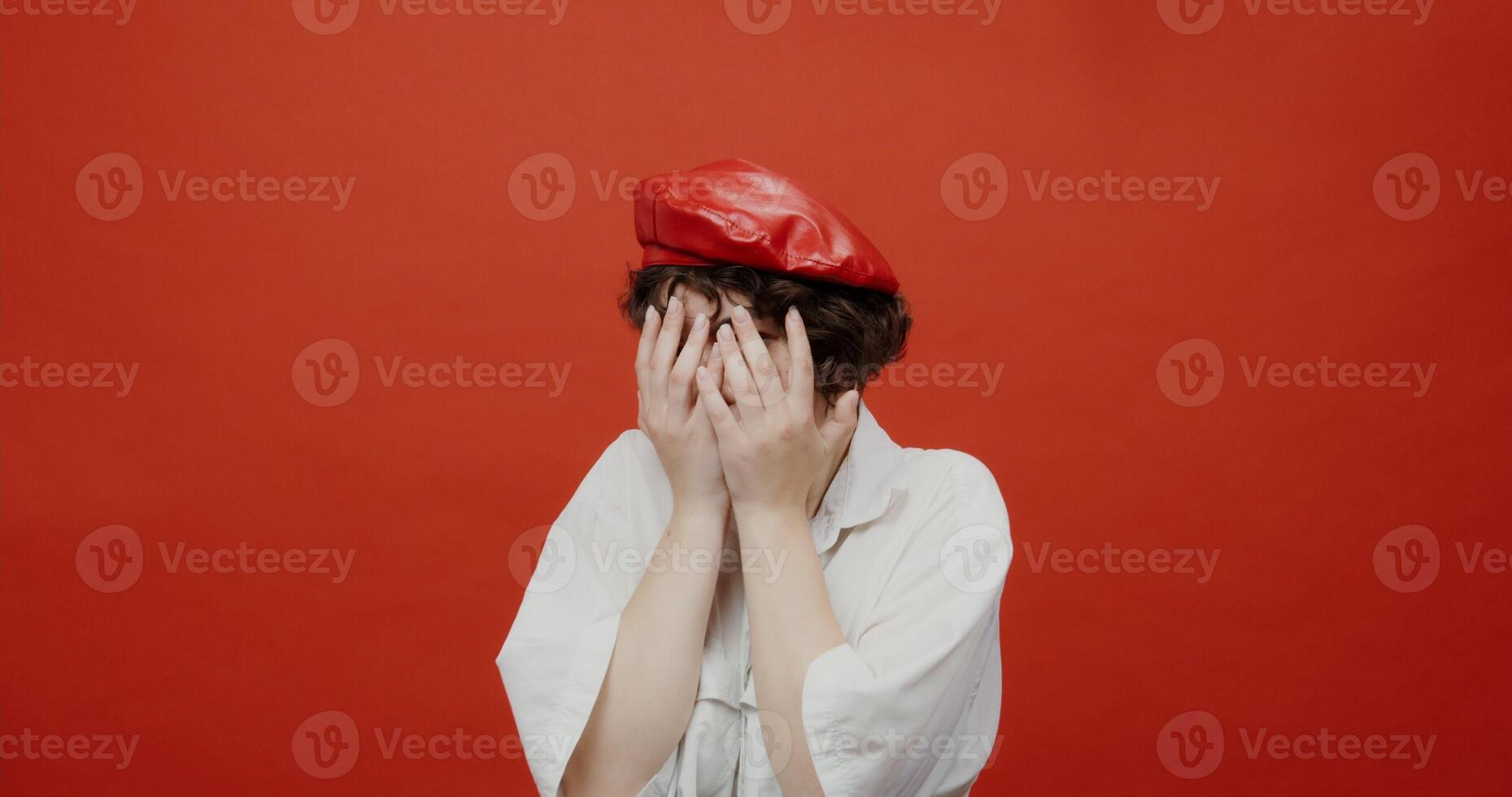 A brunette woman of European appearance with a short haircut in a red panama hat is frightened of something in front, bends over and covers her face with her hands. Shooting on a red background. photo