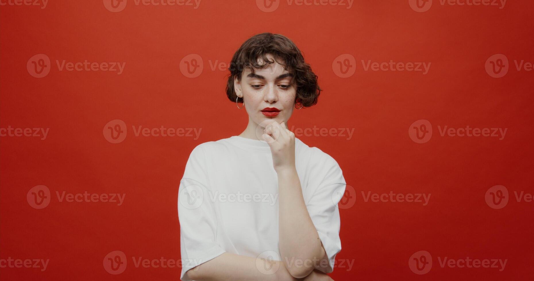 A brunette woman of European appearance with a short haircut rubs her chin in thought and rejoices at finding a solution. Shooting on a red background. photo
