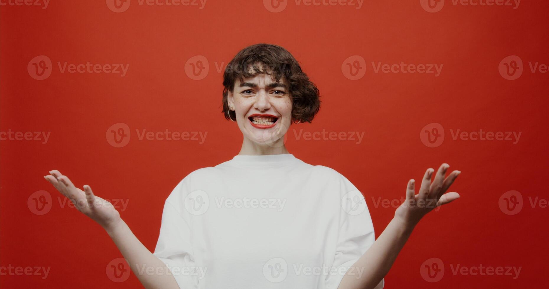A brunette of European appearance with a short haircut with red lipstick cannot understand what is happening, gets angry, then waves her hand at everything and leaves. Shooting on a red background. photo