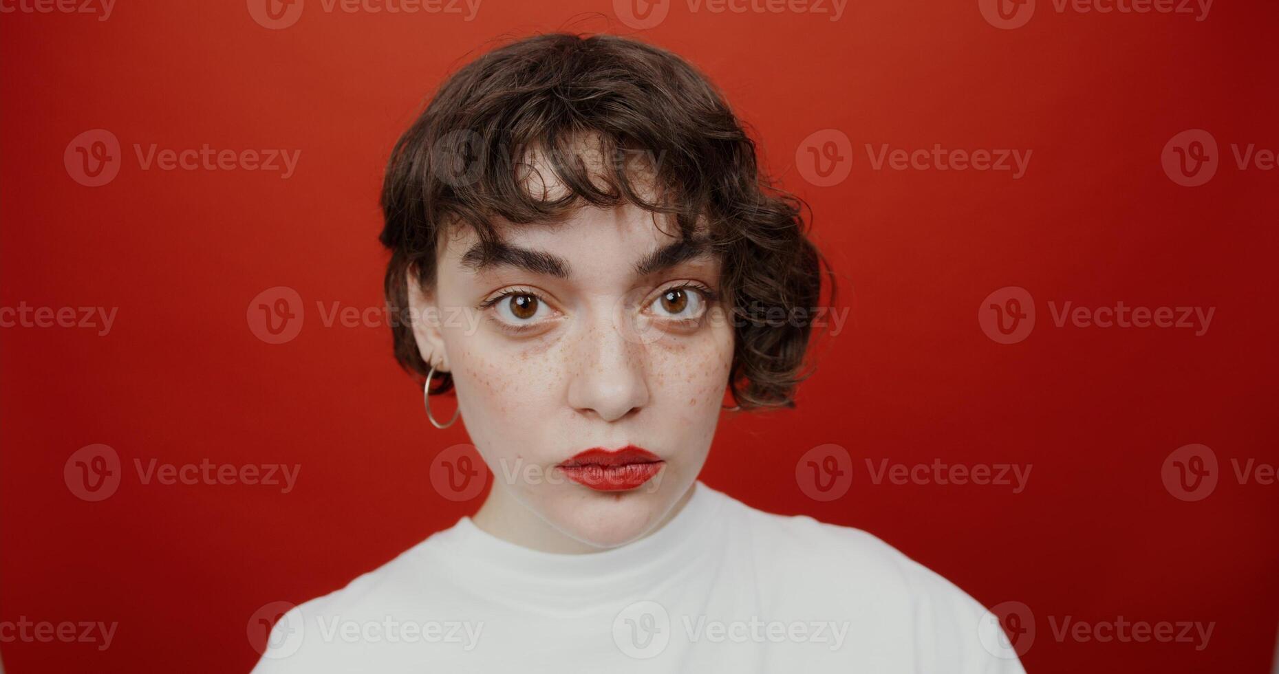 A short-haired brunette of European appearance with red lipstick is staring straight into the camera while chewing gum. Shooting on a red background. photo