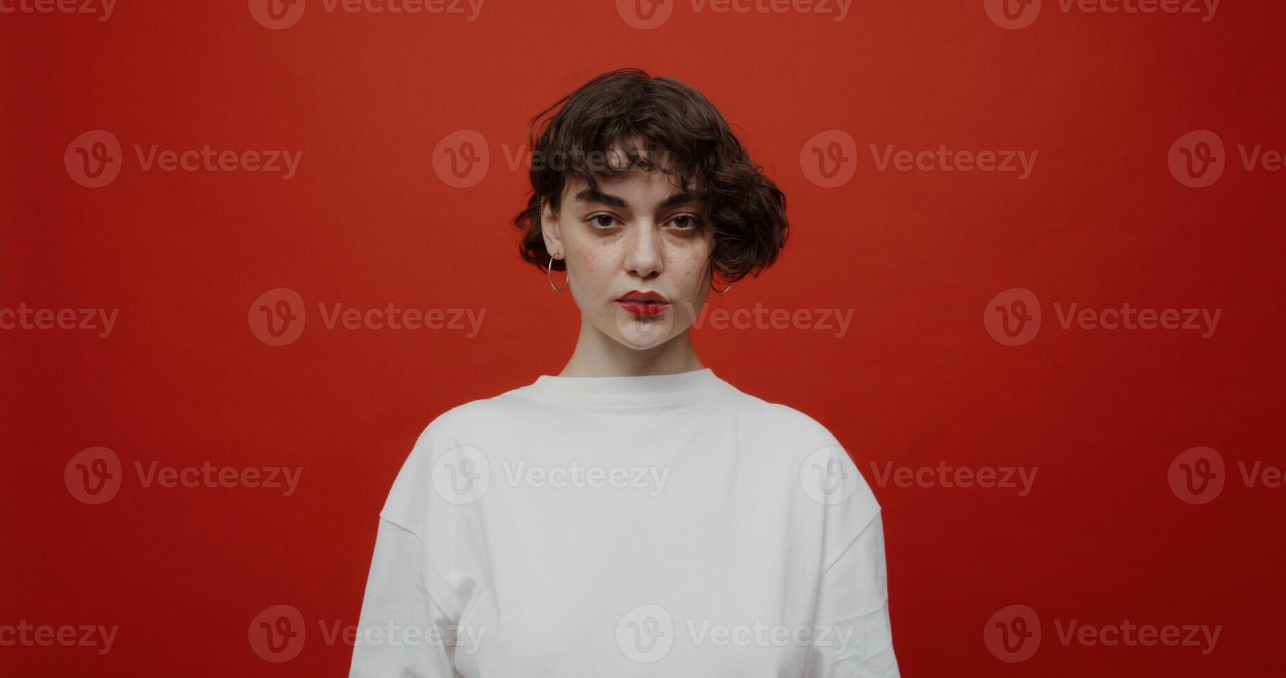 A brunette of European appearance with a short haircut with red lipstick seriously looks at the camera, putting on a black medical mask. Shooting on a red background. photo