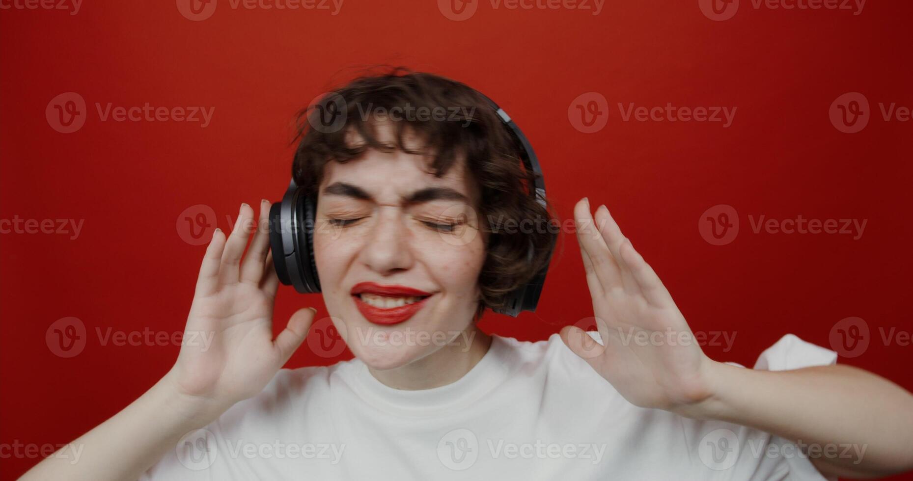 A brunette of European appearance with a short haircut with red lipstick in headphones is dancing rhythmically, eyes closed with pleasure. Shooting on a red background. photo
