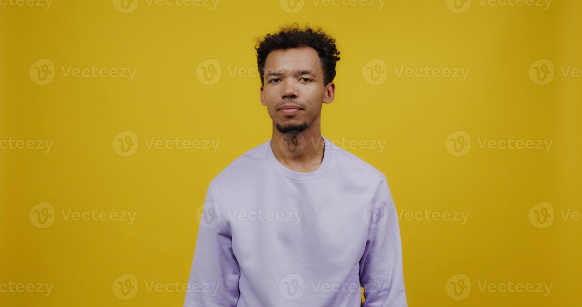 Young mulatto man with black curly hair waves his hand hello and smiles while looking at the camera. Shooting against a solid yellow background. photo