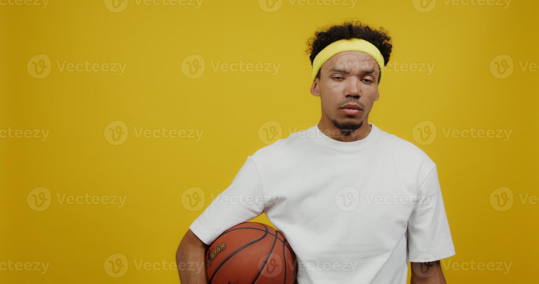 A young mulatto man with black curly hair with a bandage on his head looks seriously at the camera, clutching the ball with his hand. Shooting against a solid yellow background. photo