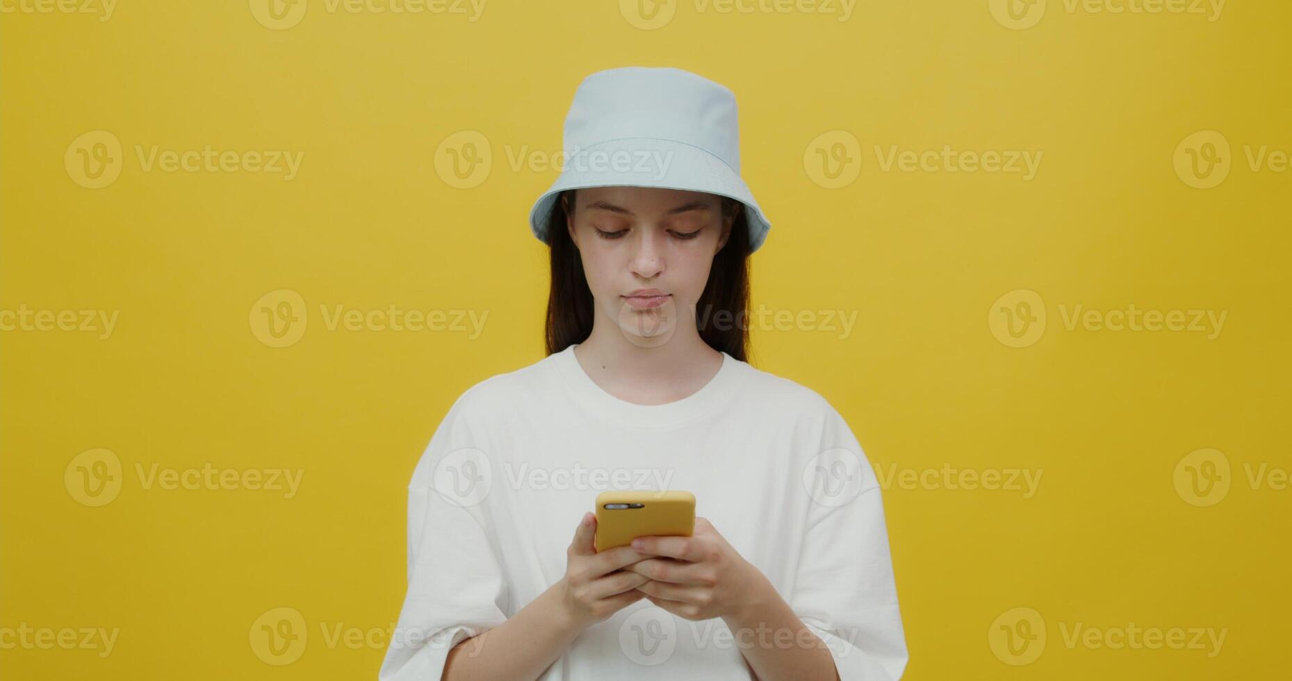 A beautiful young woman of European appearance in a panama hat smiling, chatting on a mobile phone. Portrait videography with a solid yellow background photo