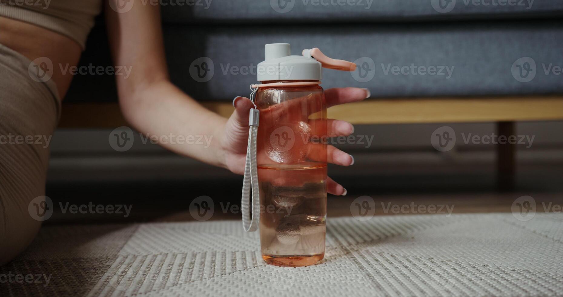 A girl in sportswear with fair skin sits on the floor and takes a sports water bottle standing on the floor photo